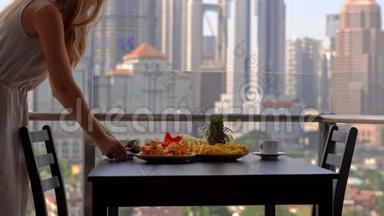 年轻的女家庭教师在阳台上准备早餐，俯瞰市中心的摩天大楼