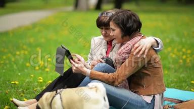 春天在公园野餐时和小狗一家人快乐。 成年女儿和她的母亲正在户外使用平板电脑。