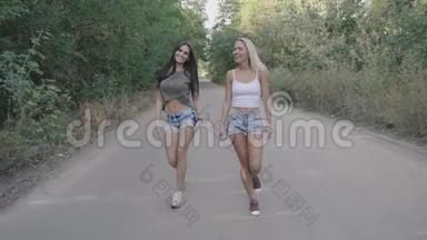 两个热辣的女人一起在森林或公园的路上散步。 金发美女和黑发美女沿着这条路走。 Stadicam