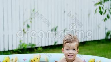一个四岁的男孩躺在一个孩子`充气游泳池里，溅起水花，微笑着。 附近的漂浮玩具。 在花园里，在