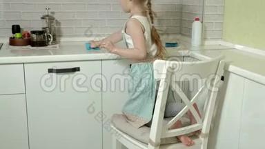穿着<strong>围裙</strong>的漂亮小<strong>女孩</strong>站在椅子上擦拭桌子，明亮的厨房里的工作表面
