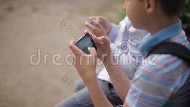 两个男孩坐在长凳上玩网络游戏。 一个带背包的男孩。 年轻人用电话