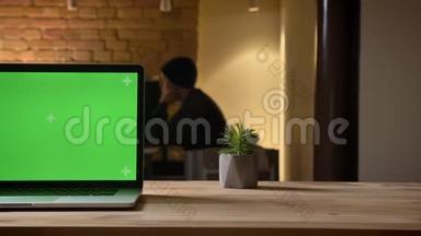 通过相机拍摄笔记本电脑的特写镜头，办公室的办公桌上有绿色屏幕，员工在室内