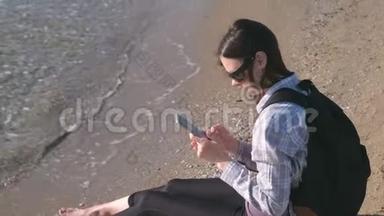 旅行者女孩带着背包坐在沙滩上，在手机上输入一条信息。