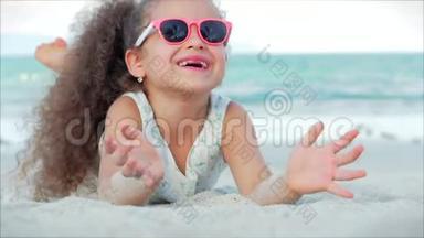 粉色眼镜中美丽的小女孩的特写镜头，可爱的微笑看着镜头，躺在沙滩上