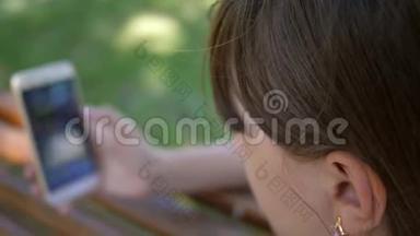一个使用智能手机的年轻女孩正在公园的一家网上商店里的长凳上翻页。 年轻的千禧年