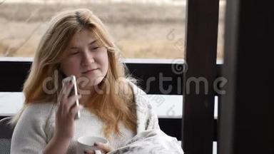舒适的家居概念。 年轻女子坐在格子花格里的露天露台上，手里拿着一杯咖啡在电话中交谈