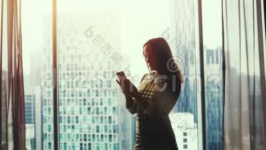 美丽的年轻女子站在窗边，在美丽的夕阳和模糊的城市背景下使用智能手机。