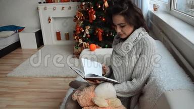 漂亮的女孩坐在沙发上的圣诞树旁，拿着记事本，望着窗外