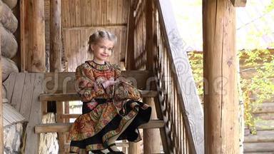 穿着传统服装的小漂亮女孩坐在一座古老的乡村木屋前台阶上的肖像。 全额