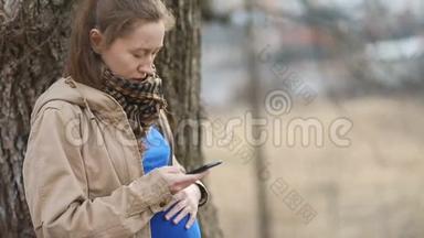 年轻的孕妇穿着浅棕色的外套和<strong>蓝色</strong>的t恤，深情地用手轻轻地<strong>滚动</strong>着智能手机
