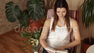 一个年轻漂亮的女孩从桌子上拿起一本书开始阅读。 坐在椅子上看书的女人。