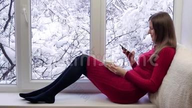 穿着红裙子的漂亮女孩坐在窗台上，喝咖啡，用智能手机。 冬天在外面。
