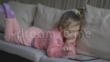 漂亮的小女孩躺在沙发上用平板电脑。 小孩子在家里的橙色沙发上玩笔记本电脑。 一点点