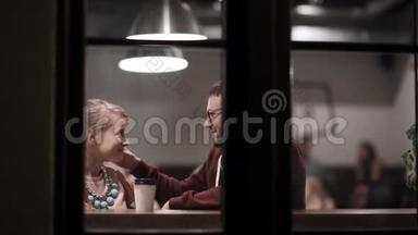 通过窗口查看.. 年轻夫妇坐在咖啡馆里，喝咖啡，聊天，晚上一起玩。