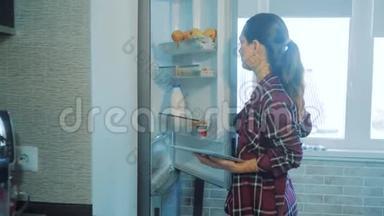 女孩从冰箱里拿食物，看着一台数字平板电脑。 智能家居概念女孩慢动作视频。 妇女