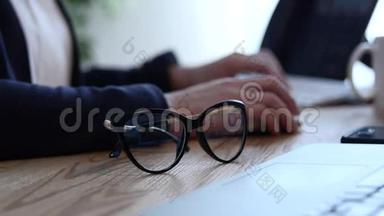 办公室现场有笔记和试卷，一支笔和一副眼镜.. 一个年轻的女人`她的手在电脑上工作