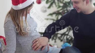 圣诞老人帽里快乐的年轻家庭。 丈夫摸着肚子，他怀孕的妻子靠近美丽的装饰圣诞树。 慢慢慢慢