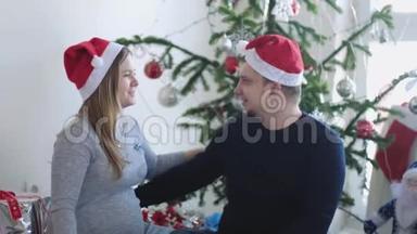圣诞老人帽里快乐的年轻家庭。 丈夫在美丽的圣诞树旁拥抱他怀孕的妻子。 慢动作