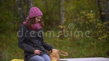 年轻漂亮的女人从包里拿着手机，坐在秋天公园的长凳上。 女人用智能手机