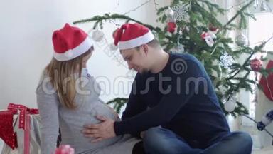 圣诞老人帽里快乐的年轻家庭。 丈夫摸着肚子，他怀孕的妻子靠近美丽的<strong>装饰</strong>圣诞树。 慢慢慢慢