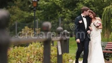 婚纱拍摄的美丽幸福夫妇微笑着，轻轻地亲吻，沿着绿色盛开的花园散步。