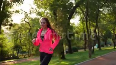 年轻的女人，长发，穿着明亮的粉红色夹克，跑在阳光明媚的<strong>城市</strong>公园里，<strong>看</strong>着<strong>镜头</strong>。 Stadicam