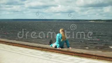 年轻的漂亮女人坐在海边的木墩上，看着这座城市。 可爱的时髦女郎带着背包