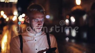 年轻迷人的男人用智能<strong>手机</strong>走在夜城的街道上。 交流、旅游、旅游、<strong>约会</strong>、商务