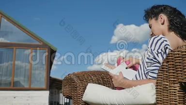 迷人的女人在柳条椅上看书，在乡间别墅的后院放松