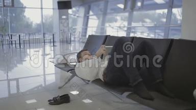 商人躺在机场候机大厅的座位上，用平板电脑