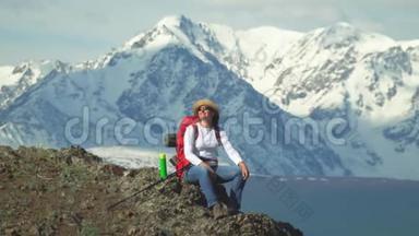 旅行者坐在山顶的女人，从热水瓶里喝热茶。 红色背包，橙色夹克。 草帽。 筷子