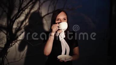 女人在黑暗的地方喝咖啡。 年轻的梦幻女子一边喝咖啡一边站在光<strong>线下</strong>