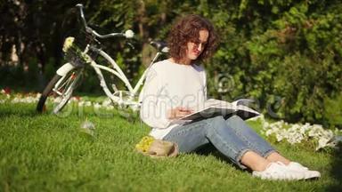 穿着白色衬衫和蓝色牛仔裤的漂亮女人正在阳光明媚的温暖日子里<strong>坐在</strong>公园的草<strong>地上</strong>看书。 她是