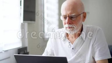 穿着劳累过度，穿着白色衣服的老人在家里用笔记本电脑工作。 揉揉他疼痛的眼睛
