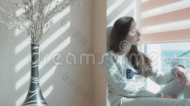 一个年轻漂亮的女人坐在俯瞰大海的窗户上。 桌子上有一个花瓶，上面有装饰花.. 光线