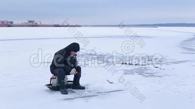 穿着深色衣服的老渔夫在河港的背景下在<strong>冰冻</strong>的河面上用冬季的鱼竿钓鱼
