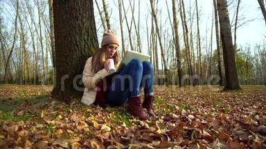 迷人的年轻女孩在秋天公园的树下，坐在地上，在笔记本电脑里喝咖啡。