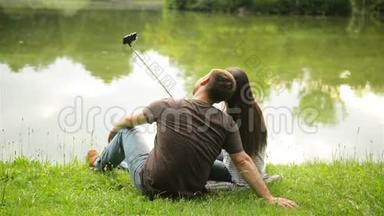 一对<strong>浪漫情侣</strong>坐在湖边绿草上用自<strong>拍</strong>棒<strong>拍</strong>照的背影。