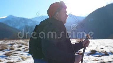 山背景下，年轻的徒步旅行者在雪地小径上用棍棒行走，在野外的侧面观看。 运动男