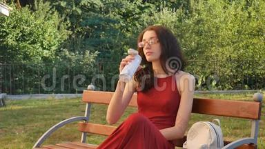 穿着<strong>深红</strong>长衣和眼镜的可爱的女青少年喝着坐在长凳上的热杯