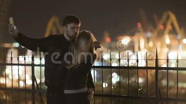 年轻的幸福夫妇在智能手机上的相机上拍摄自拍照片。 港口夜间灯光