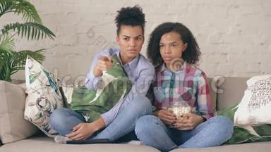 两个混血卷发女孩朋友坐在沙发上看紧张的电视节目，在家吃爆米花