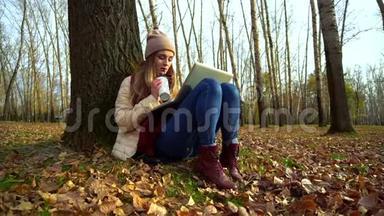 迷人的年轻女孩在秋天公园的<strong>树下</strong>，<strong>坐在</strong>地上，在笔记本电脑里喝咖啡。