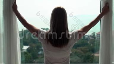 穿白色t恤的女人揭开窗帘，<strong>看着窗外</strong>。 在外面欣赏海景