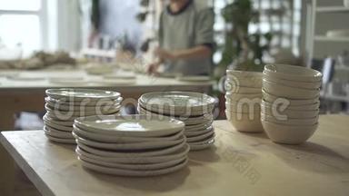 在明亮的陶瓷工作室里，<strong>一堆一堆</strong>的盘子和碗立在木桌上。