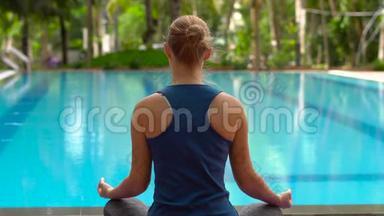 在热带的游泳池附近，年轻<strong>女子</strong>坐在<strong>瑜伽</strong>垫上以莲花的姿势练习<strong>瑜伽</strong>