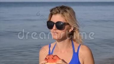 一个戴太阳镜的女人坐<strong>在</strong>海滩上<strong>吃西瓜</strong>。 一个成熟的<strong>西瓜在</strong>金发女郎手中