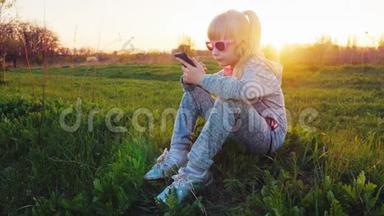 一个穿着<strong>运动服</strong>的6岁<strong>女孩</strong>坐在草地上，用手机。 日落时分