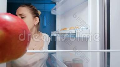 饥饿的年轻女人在冰箱里偷笑和咬苹果的4K镜头。 不健康营养概念